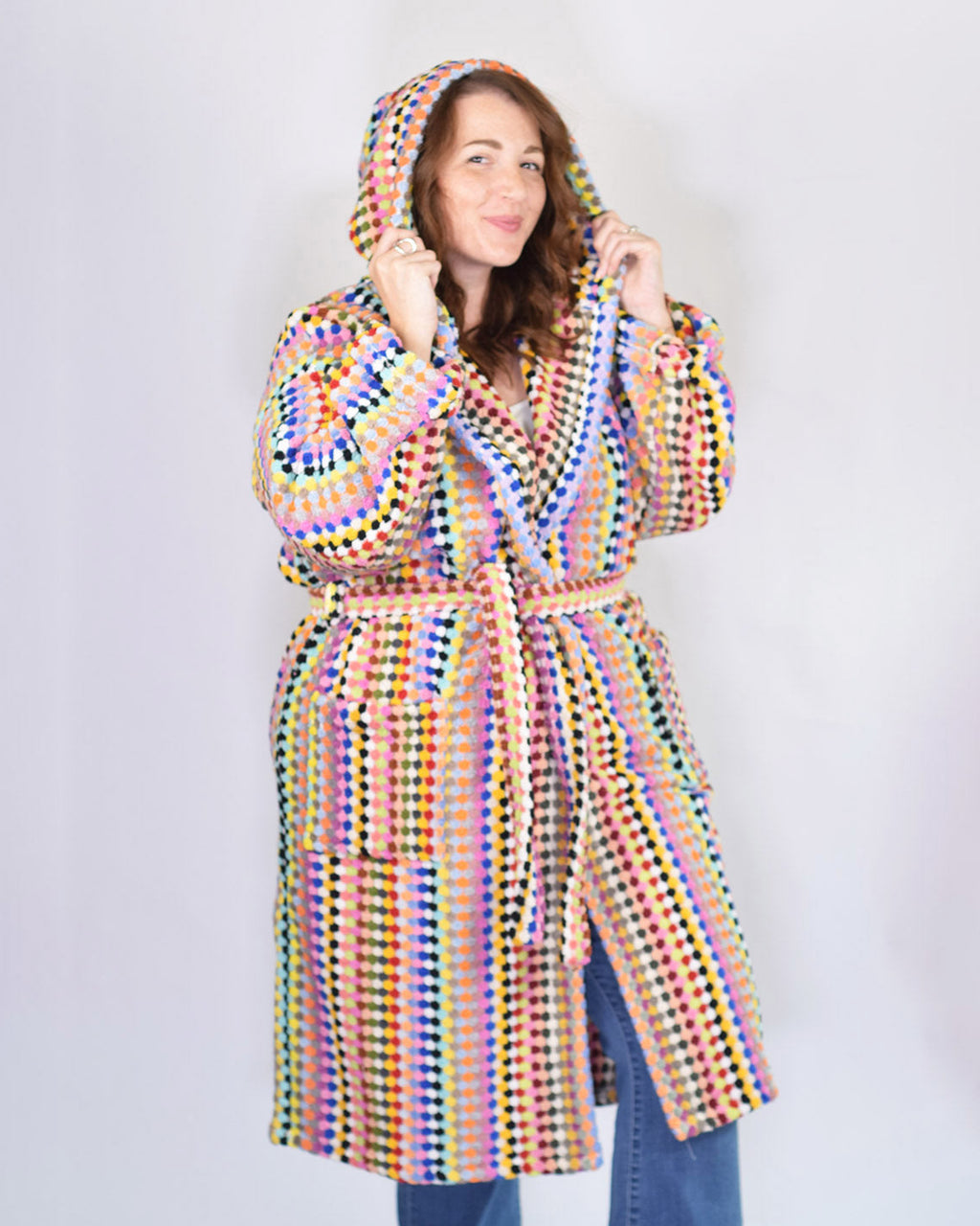 A woman wearing a hooded robe with a vibrant multicolor crochet pattern, paired with blue jeans, standing against a plain light gray background.