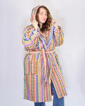 A woman wearing a hooded robe with a vibrant multicolor crochet pattern, paired with blue jeans, standing against a plain light gray background.