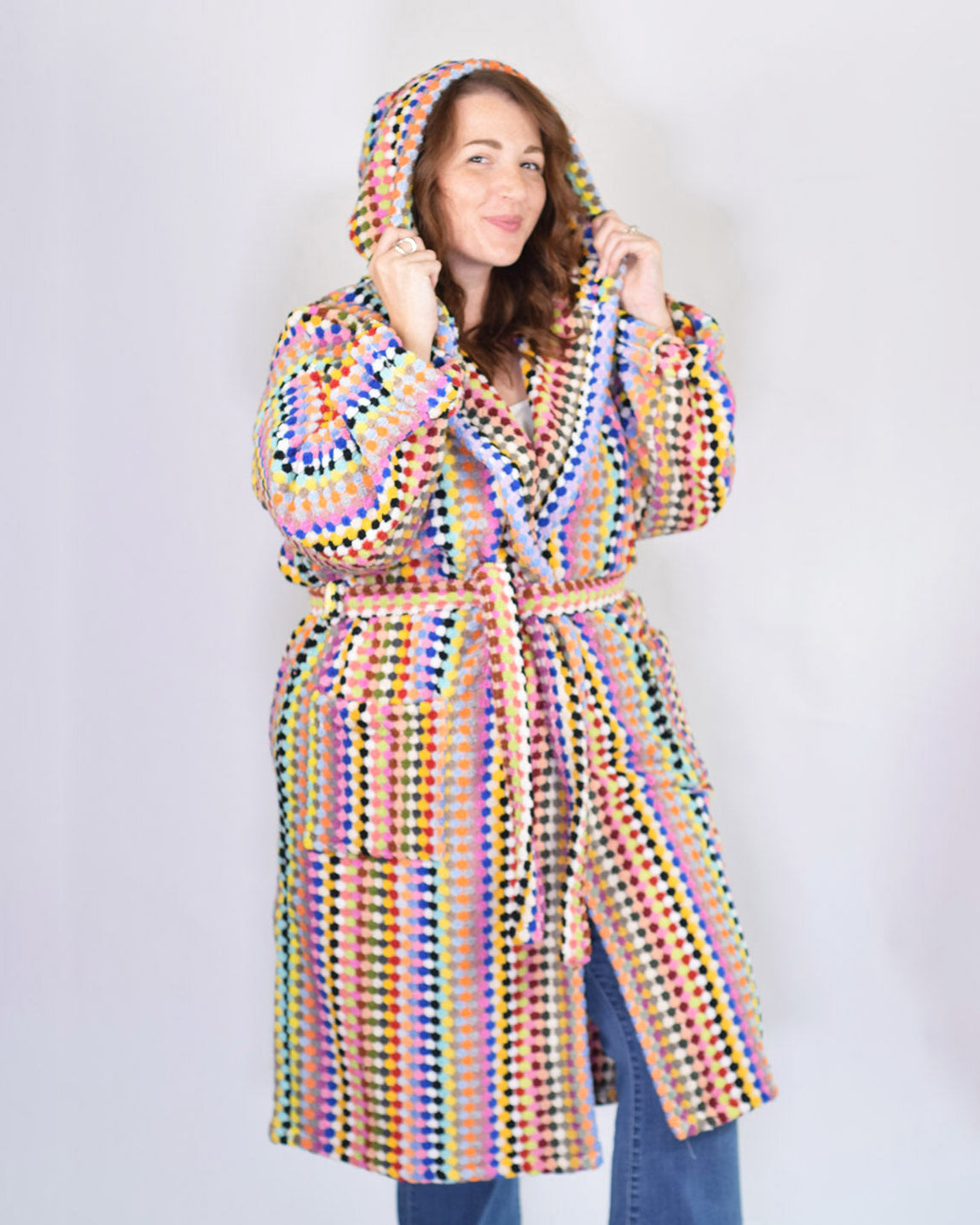 A woman wearing a hooded robe with a vibrant multicolor crochet pattern, paired with blue jeans, standing against a plain light gray background.